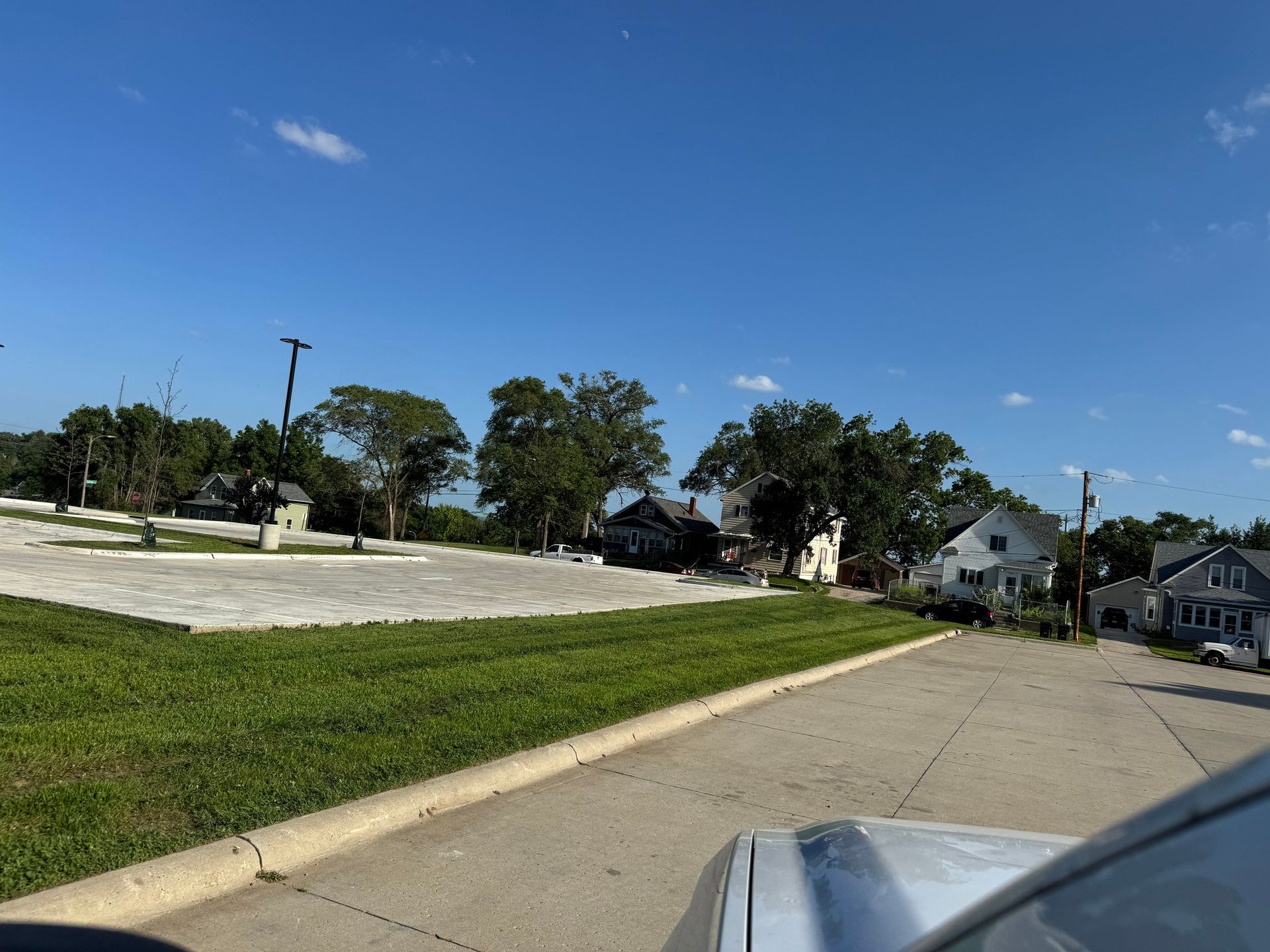 Quiet suburban street under a clear blue sky, with houses, trees, and a grassy median beside the road.