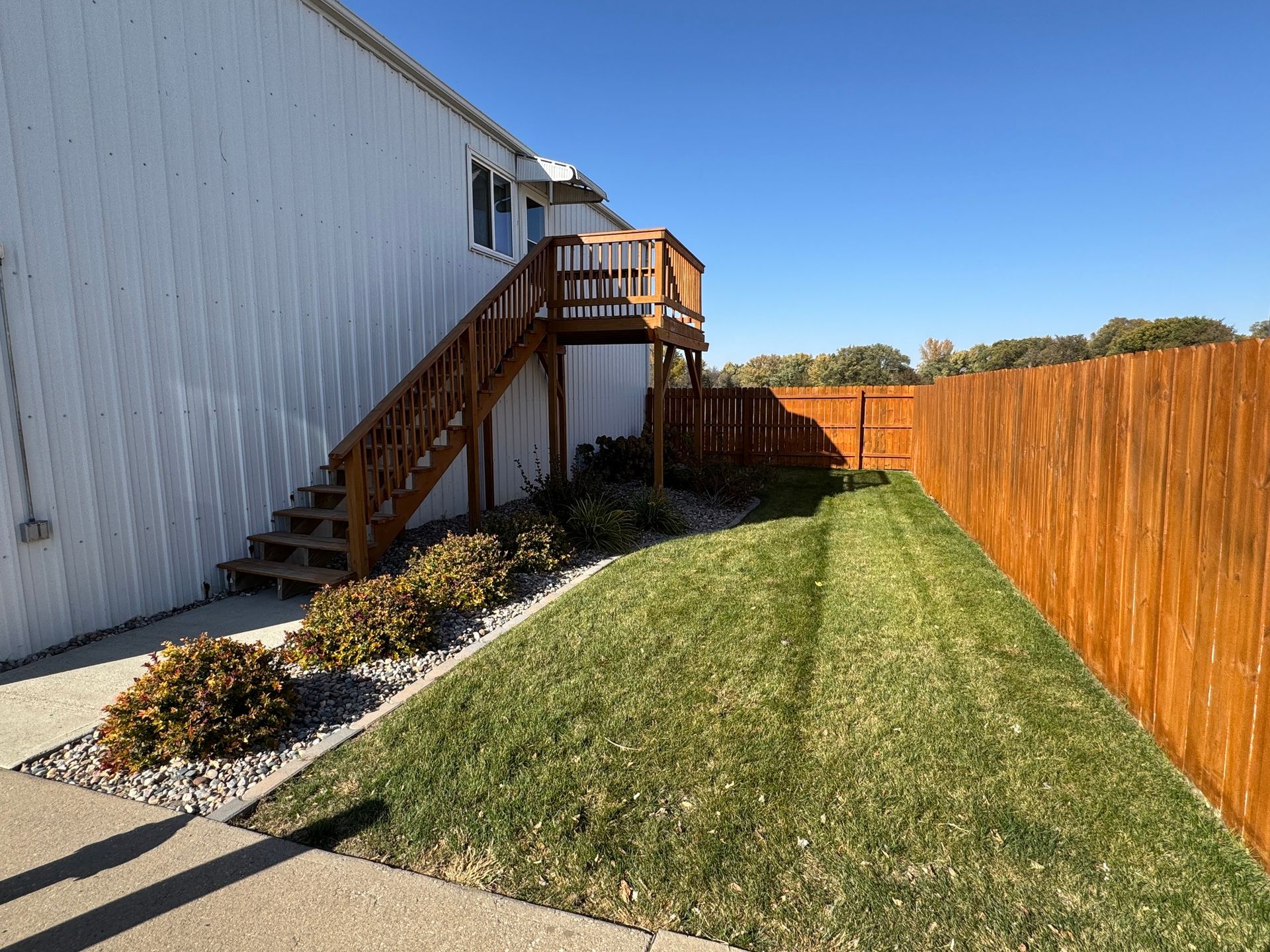 Backyard beside white building with wooden stairs, gravel path, and long grassy strip beside a wooden fence.