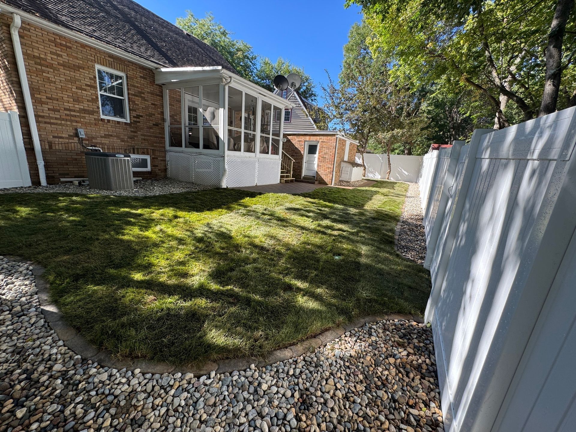 Backyard with grassy slope, pebble border, brick house, white screened porch, and tall white fence