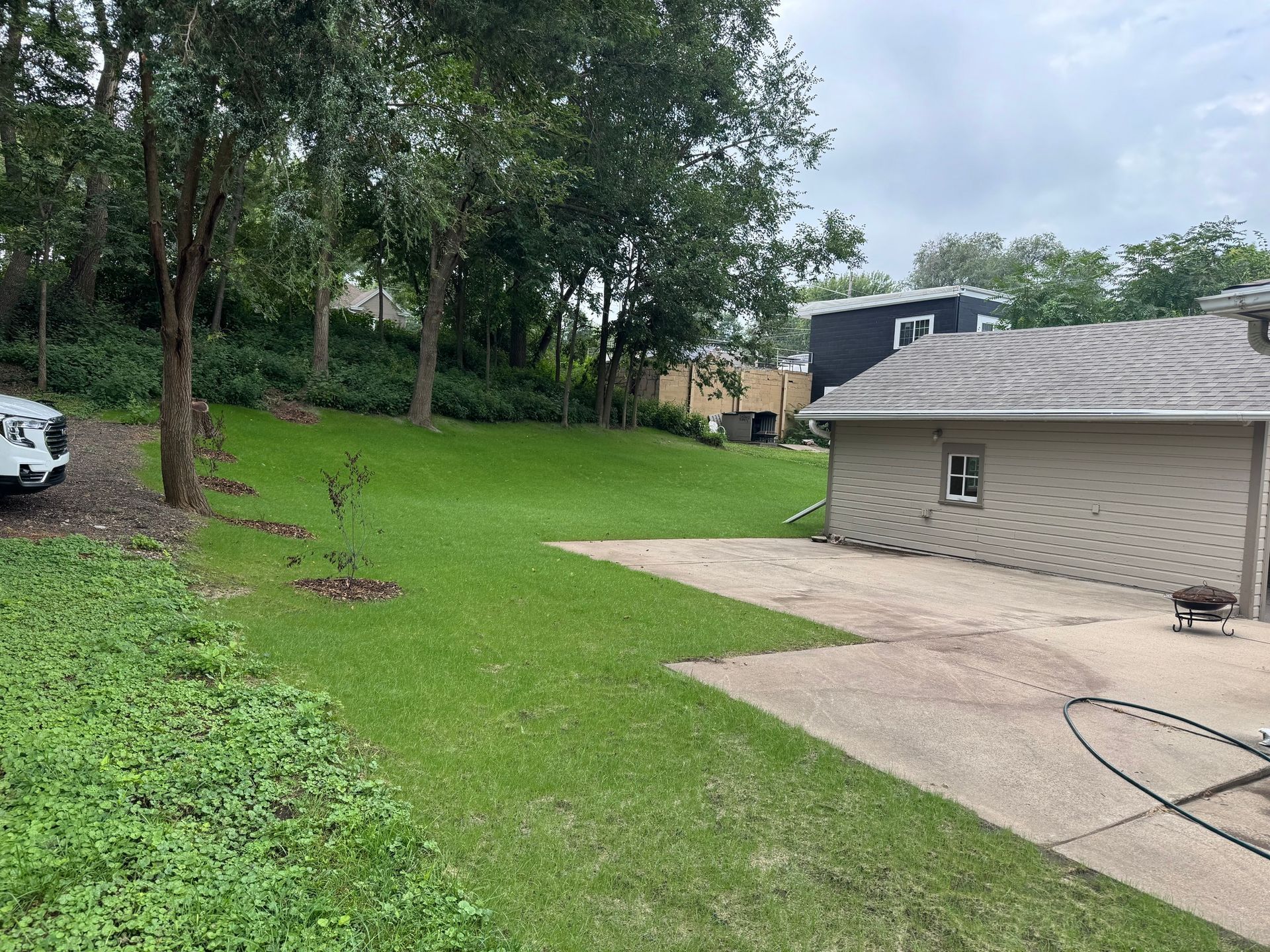 Grassy yard beside a house with a concrete driveway, trees, and a parked white car in the background