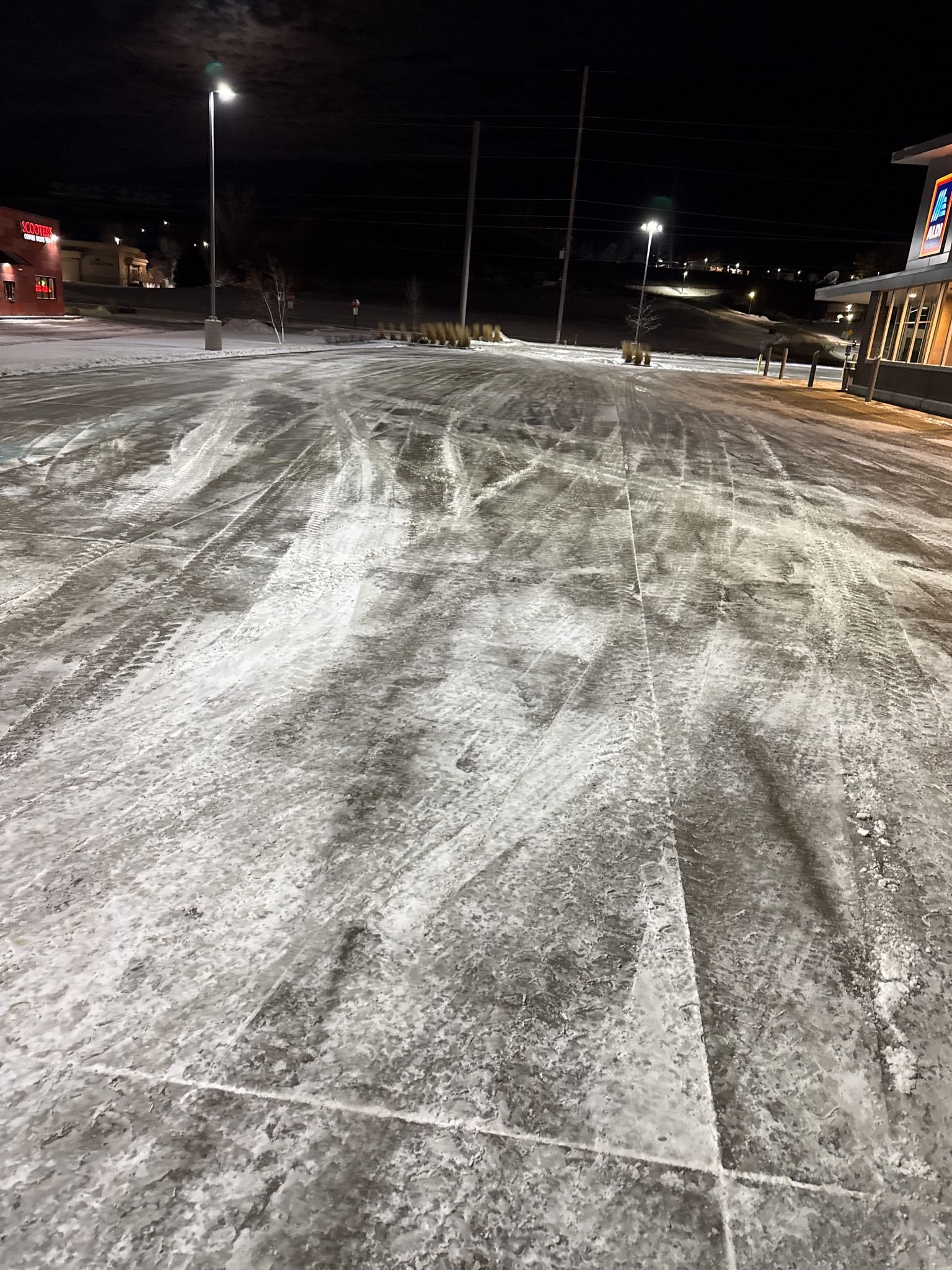 Snow-covered parking lot at night with tire tracks and storefront lights.