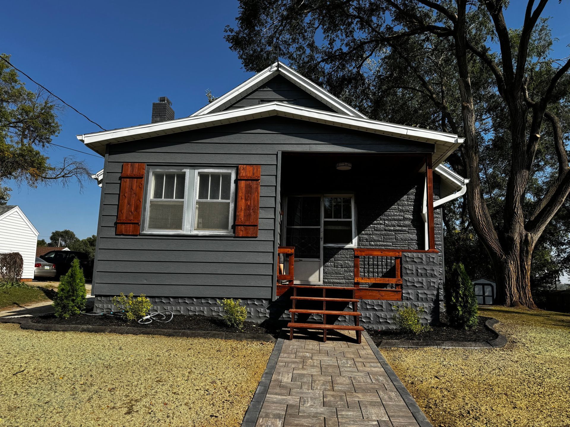Gray bungalow with red shutters, small front porch, and stone walkway under a clear blue sky