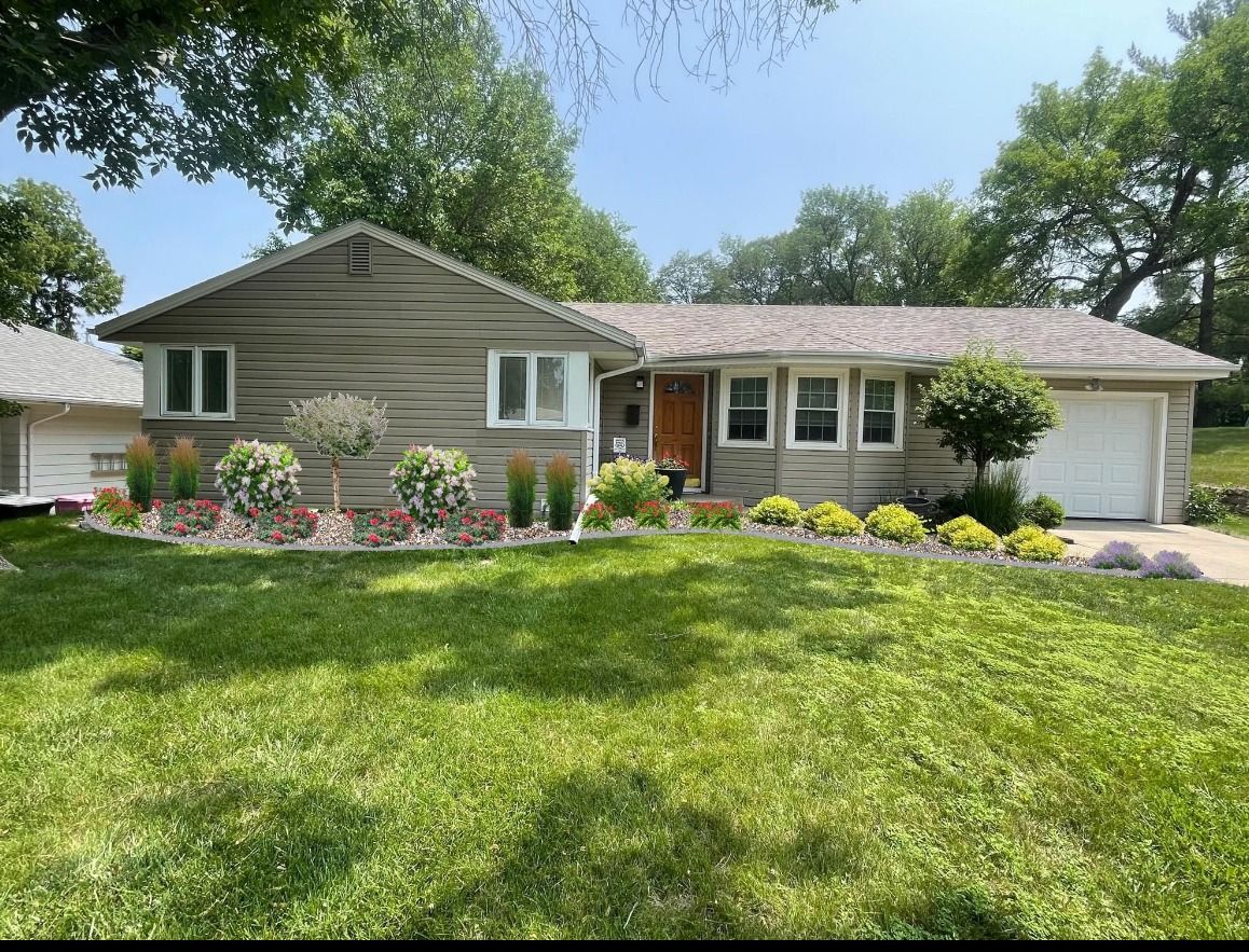 Single-story beige house with white garage, flower beds, and a bright green front lawn under a sunny sky