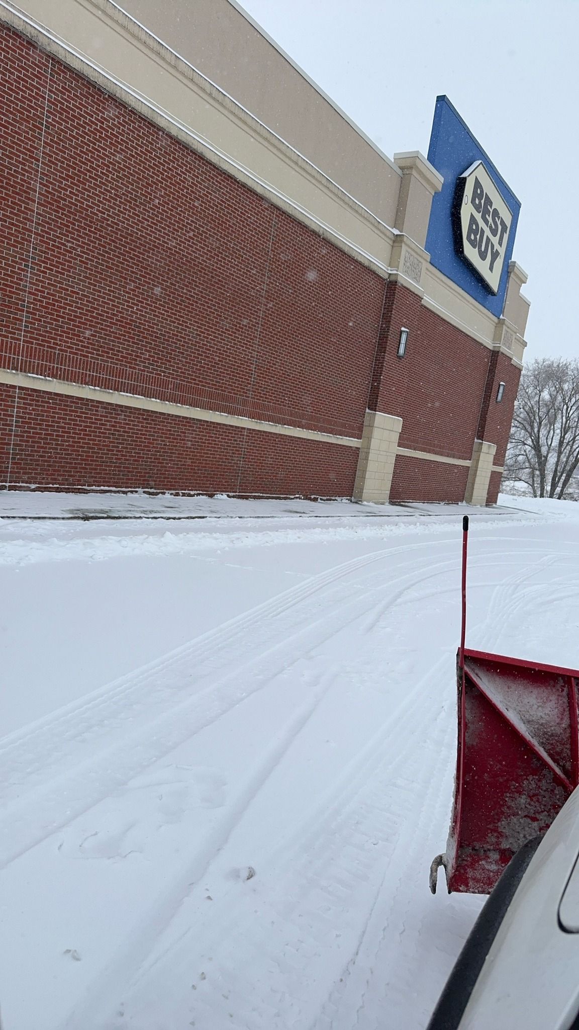 Snow-covered parking lot beside a brick store with a blue sign and a red snowplow blade in the foreground