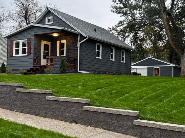 Gray house with lit front porch and detached garage on a green lawn beside a gravel driveway