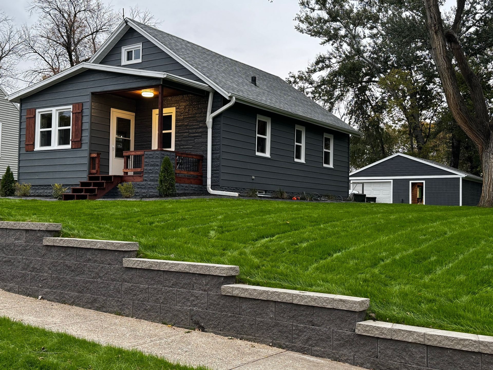 Gray house with lit porch on a grassy slope, with a detached garage in the background