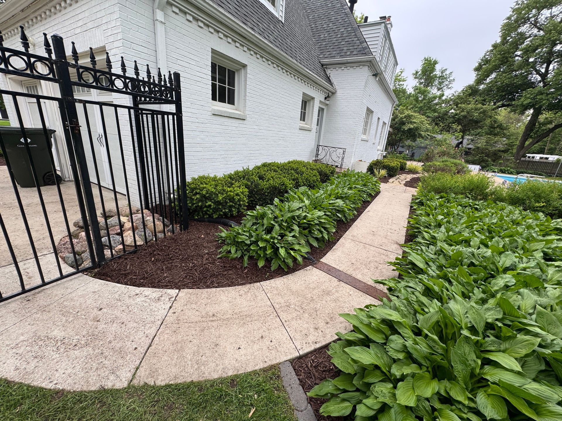 Curved garden path beside a white brick house with black iron gate and lush green shrubs