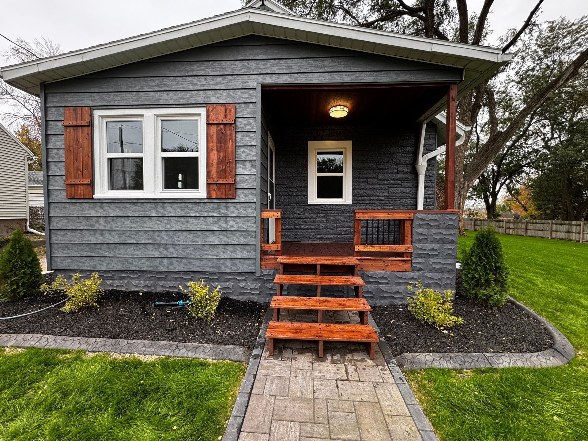 Small gray house with orange trim, front porch, and stone walkway surrounded by wet lawn and trees