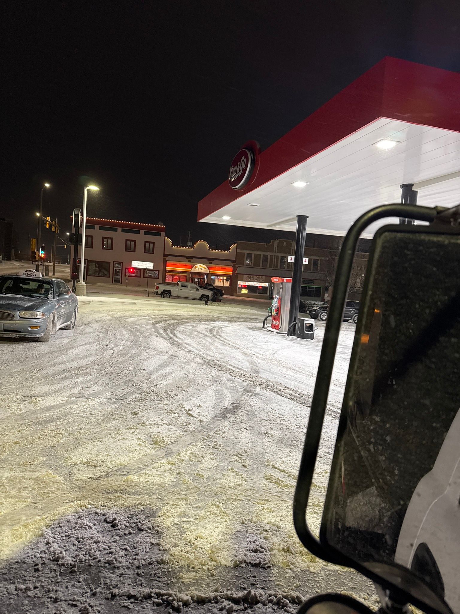 Snowy gas station at night, with illuminated pumps and parked vehicles under a red canopy.