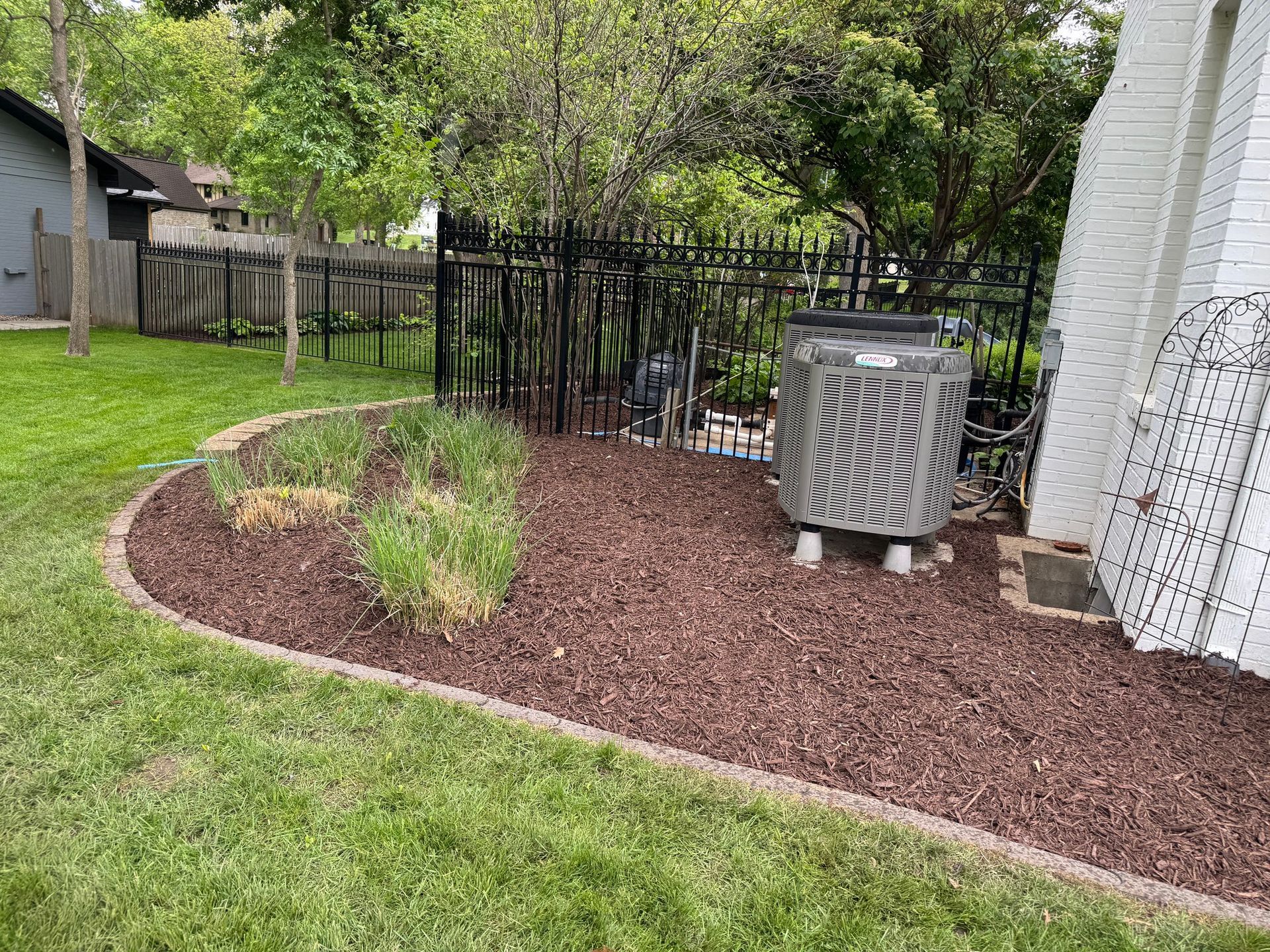 Backyard with curved mulch bed, small shrubs, black fence, and air conditioner unit near a white house