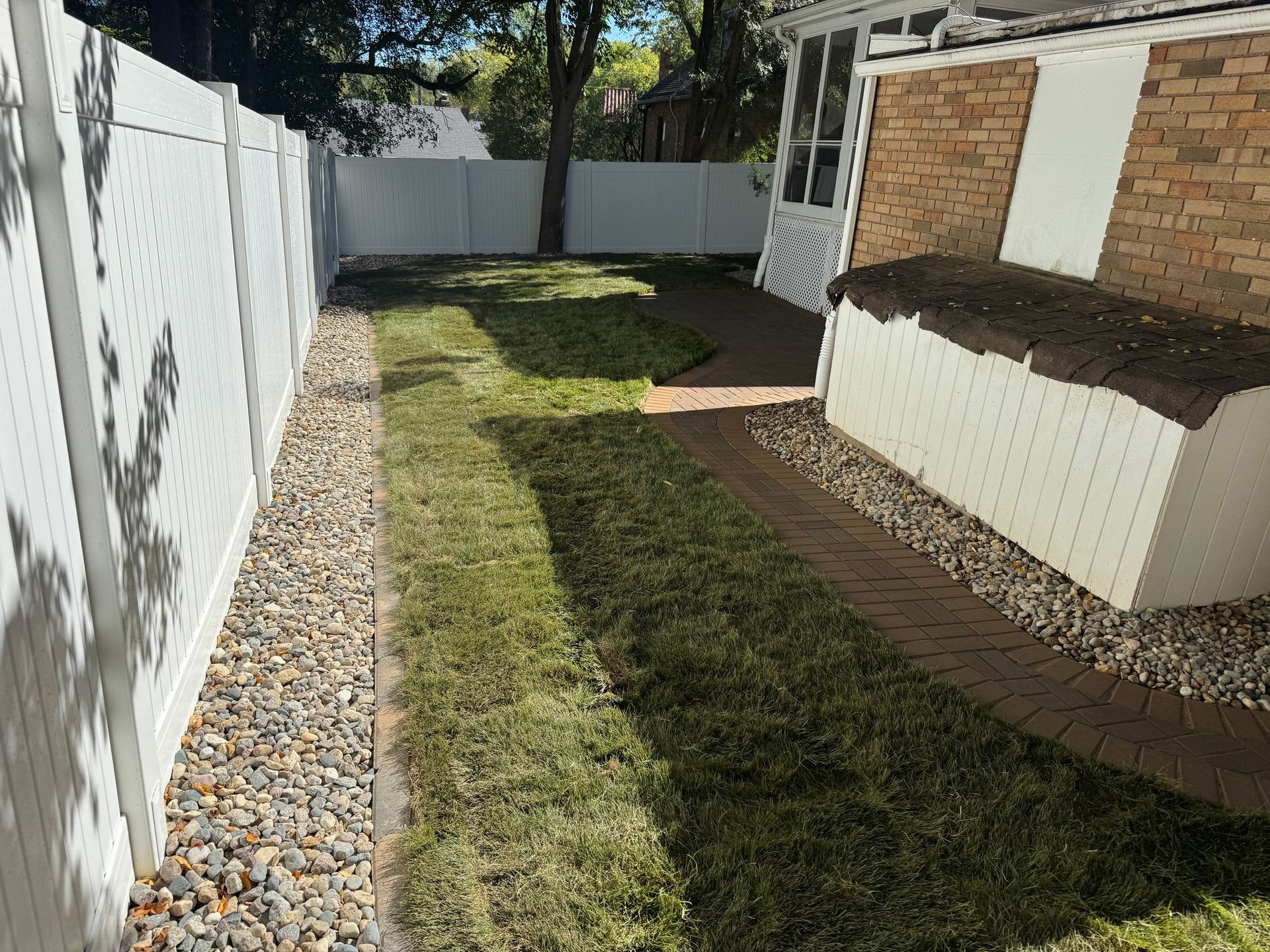 Narrow backyard with grass, brick path, white fence, and raised planter beside a house