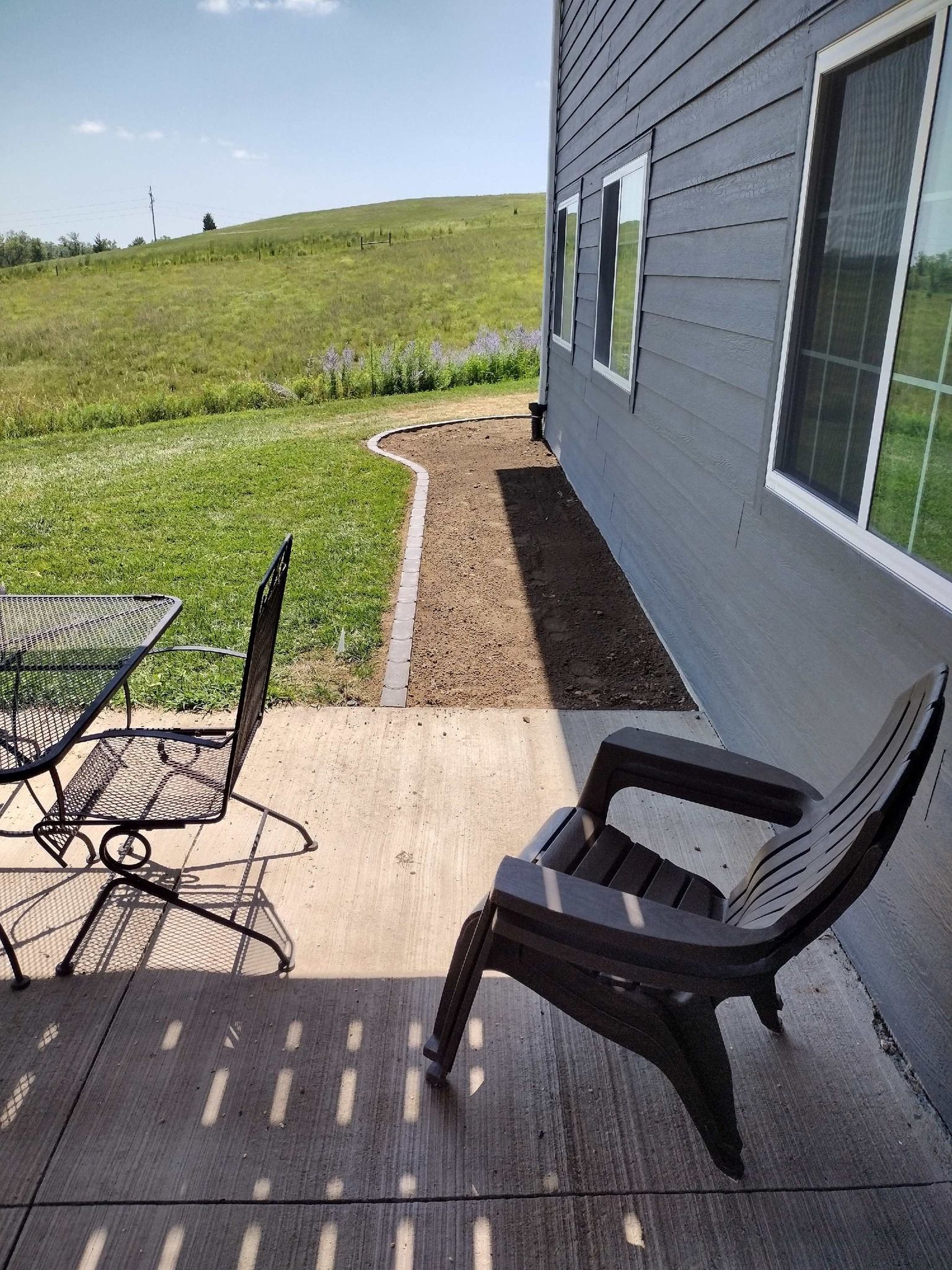Patio with black chairs beside a gray house, overlooking a grassy hill and cloudy sky