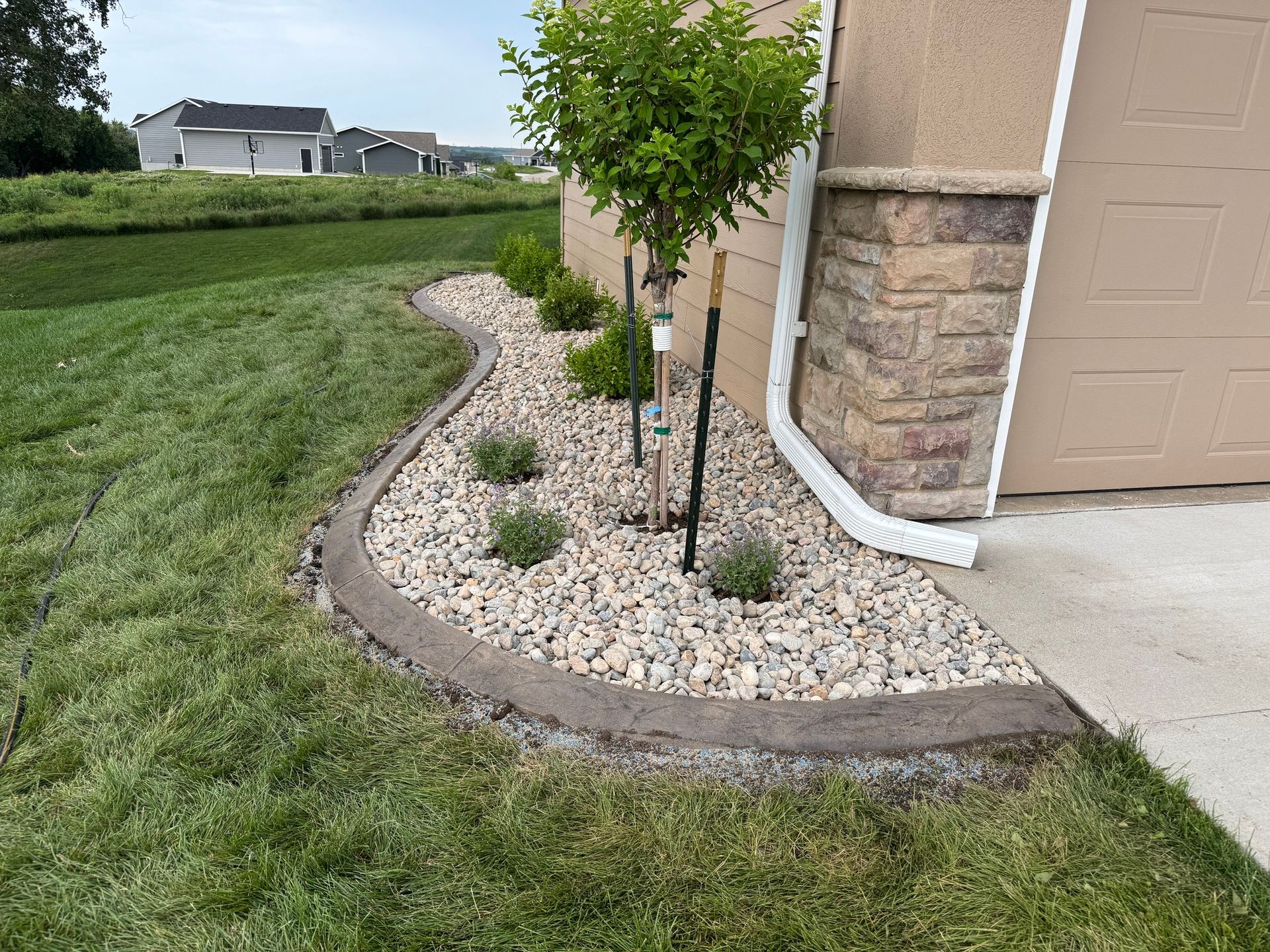 Landscaped front yard bed with rocks, small shrubs, and a young tree beside a garage and house corner