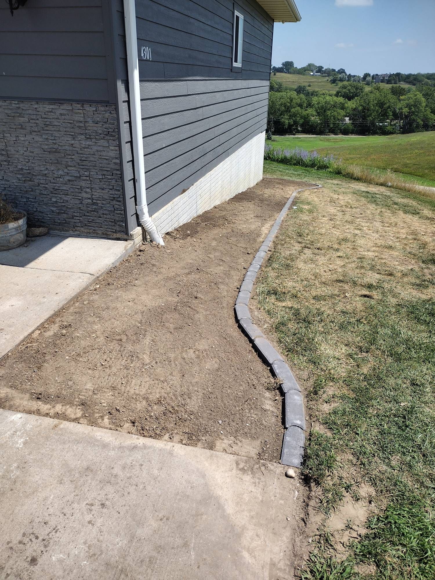 Narrow dirt strip beside gray house, bordered by concrete and grass, with a curved edging along the path