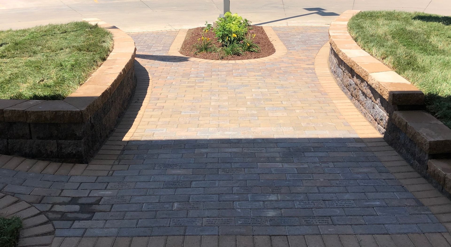 Brick walkway with steps between curved stone planters and grass leading to a paved path
