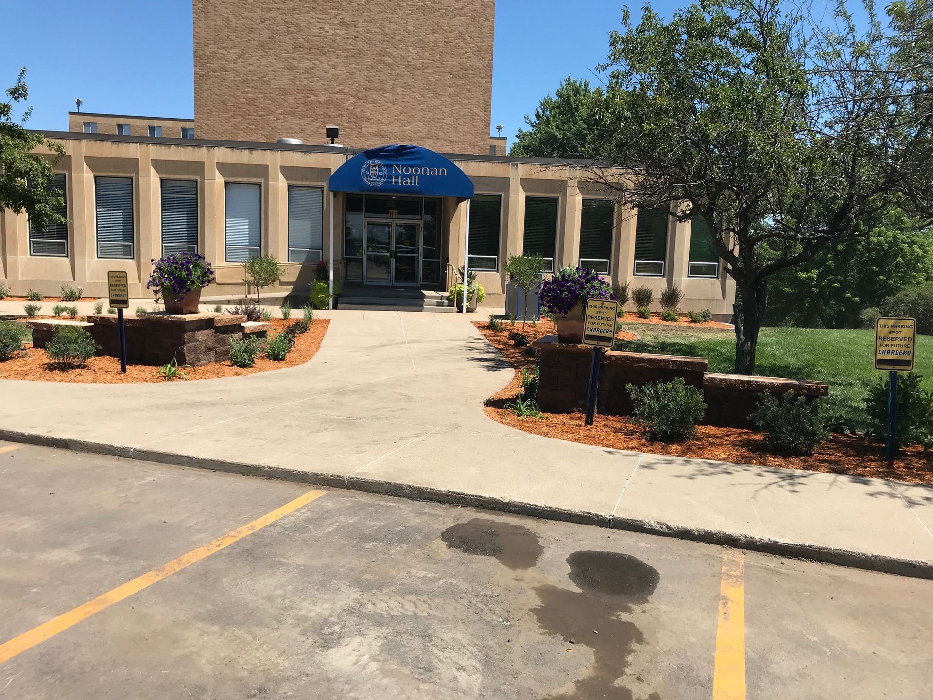Entrance to a tan brick building with blue awning, walkway, and landscaped shrubs in front