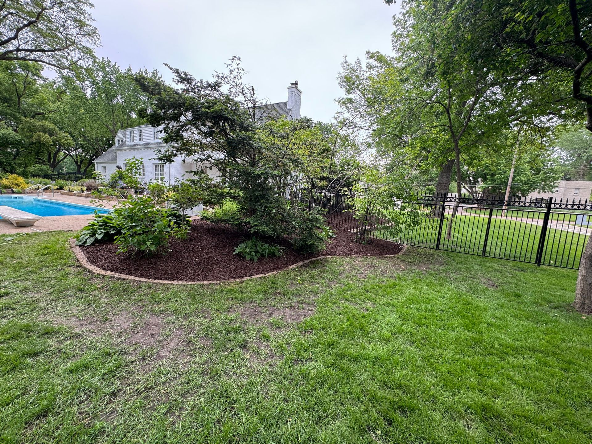 Backyard with green lawn, mulched shrub bed, fence, and above-ground pool near a white house