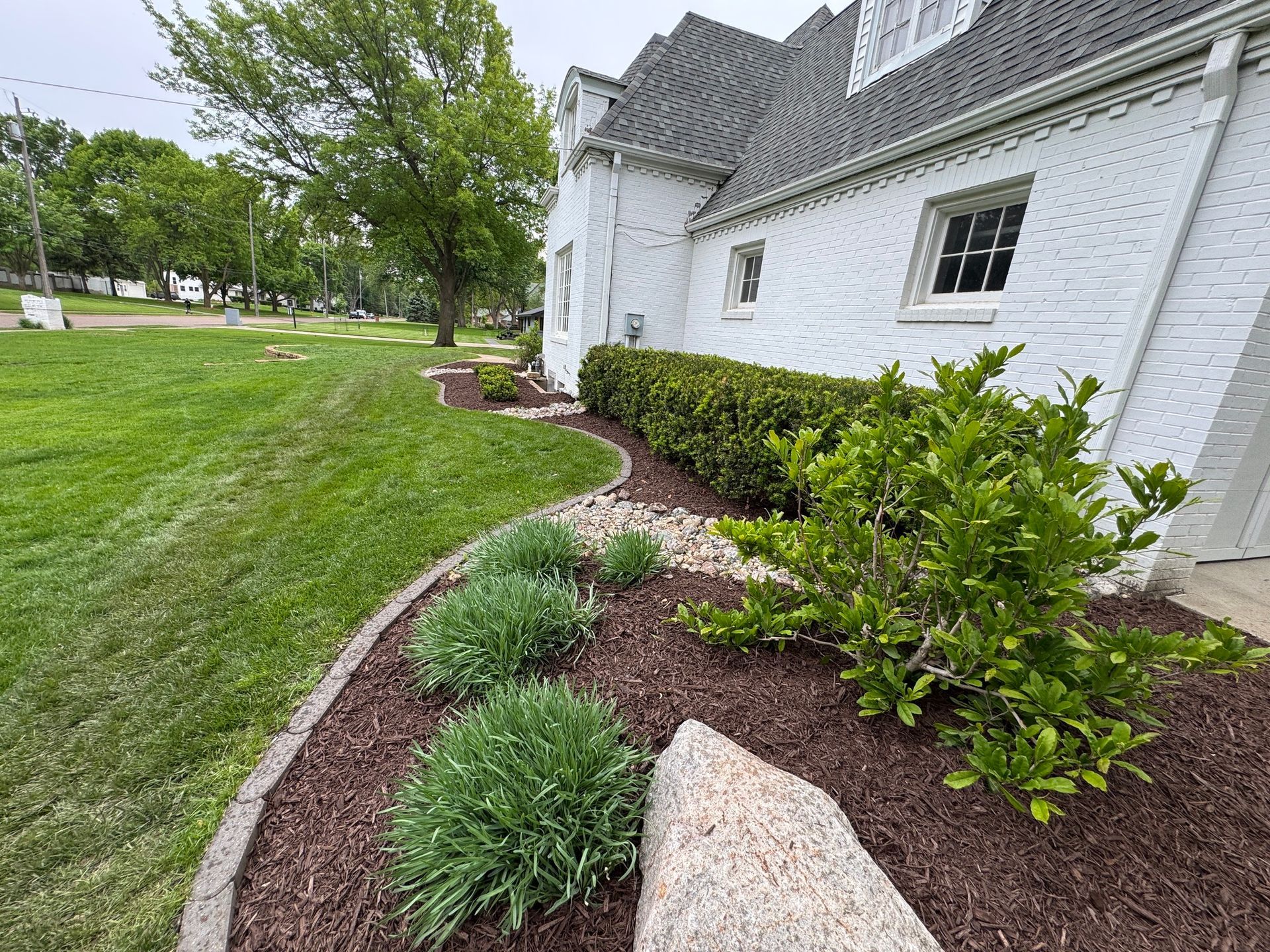 White brick house with a curved landscaped flower bed, green shrubs, and a manicured lawn