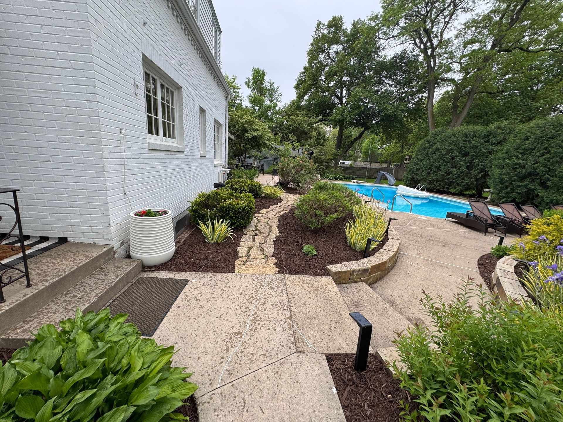 Backyard patio with white house, stone path, garden beds, and blue swimming pool