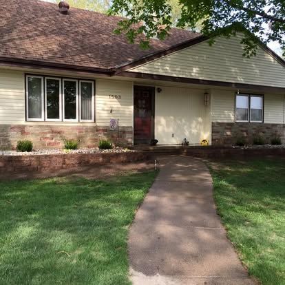 Single-story house with beige siding, brown roof, front walkway, and green lawn