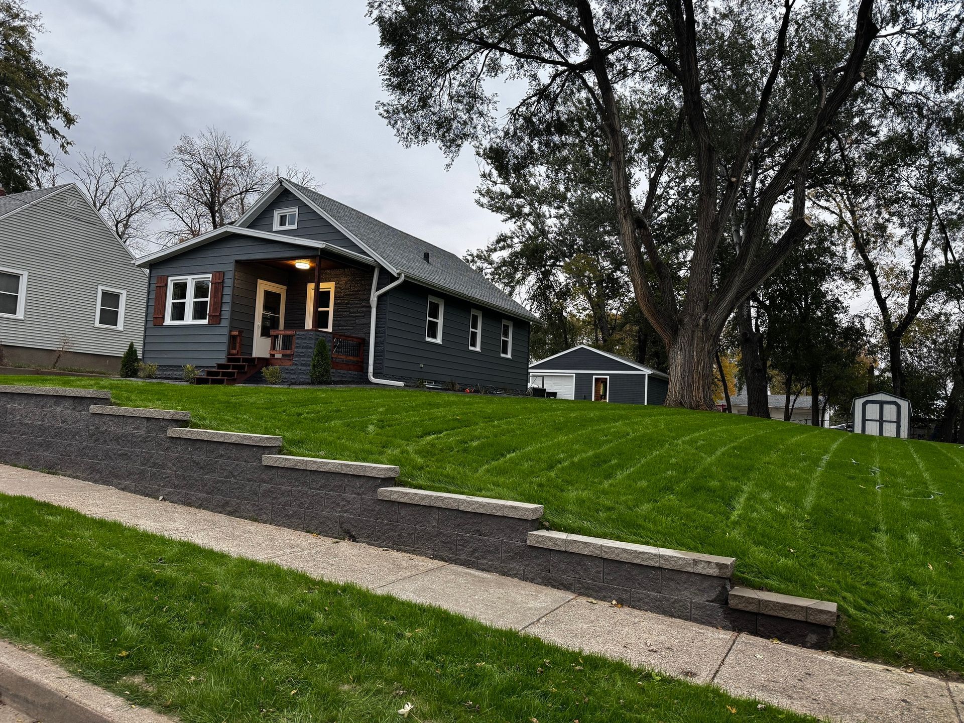 Gray house with lit porch on a sloped lawn and stone steps under cloudy skies