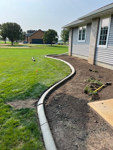Curved garden bed along a house with fresh soil edging a green lawn and sidewalk.