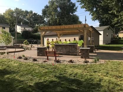 Workers in neon vests build a wooden pavilion in a park with benches and trees.