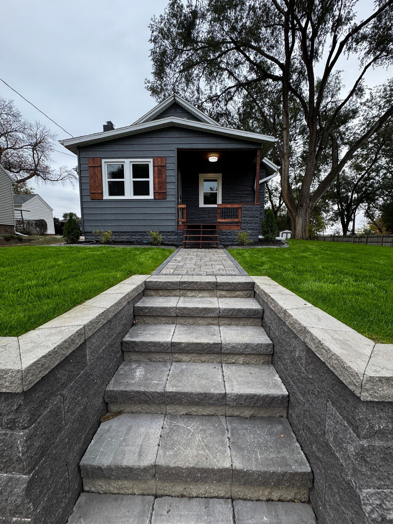 Small gray house with a front walkway and concrete steps, framed by green grass and bare trees.
