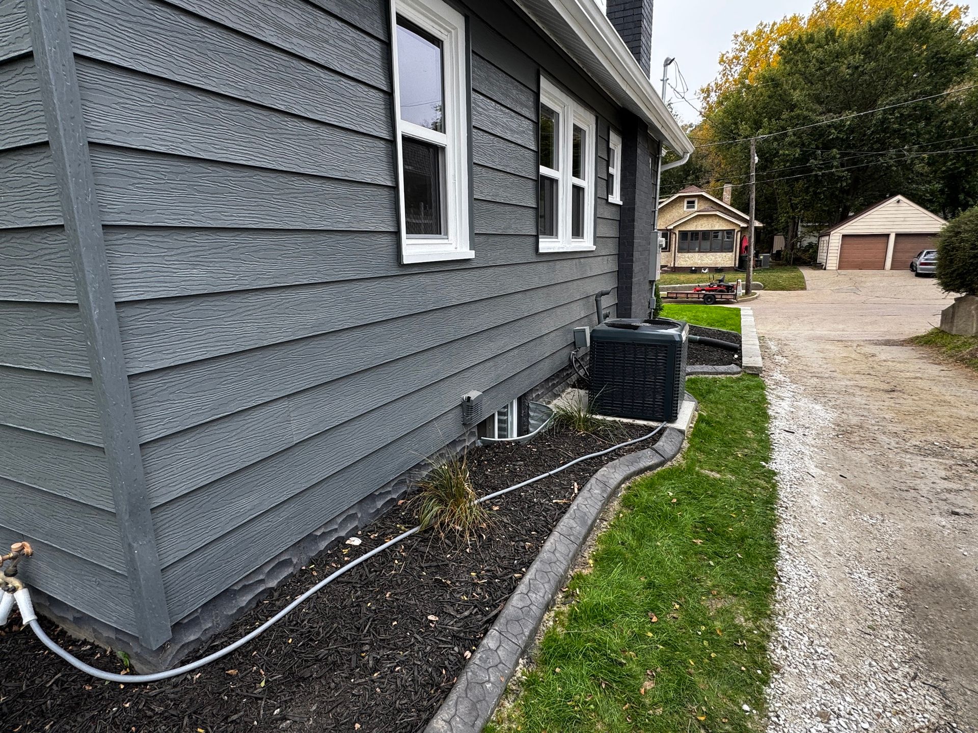 Gray house exterior with black downspout, mulch bed, and gravel driveway beside a grassy strip.