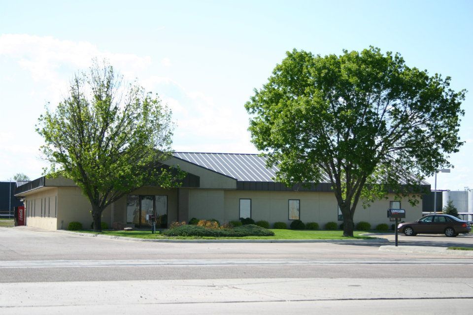 Small beige building with a metal roof beside trees and a parked pickup truck on a sunny street.