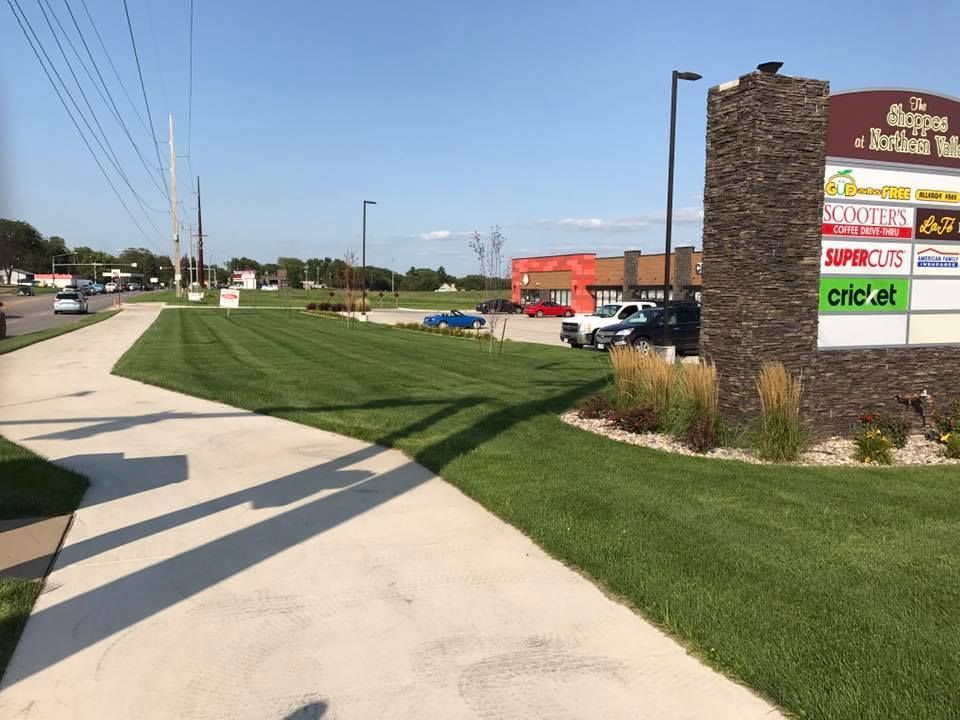 Sidewalk along a roadside strip mall with a grassy verge, sign, parked cars, and utility poles under a clear sky.