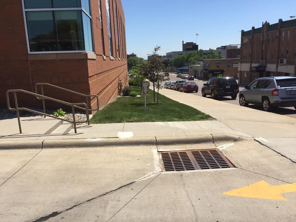 Brick building corner with sidewalk ramp, storm drain, parked cars, and street with yellow arrow marking