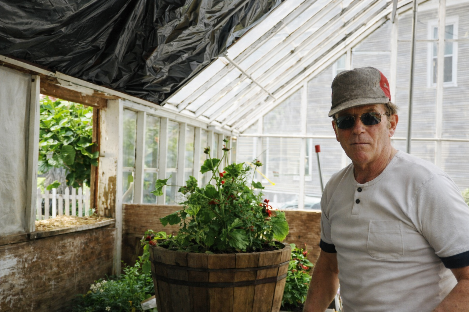 Person standing in a covered porch beside a potted plant, wearing a hat and sunglasses
