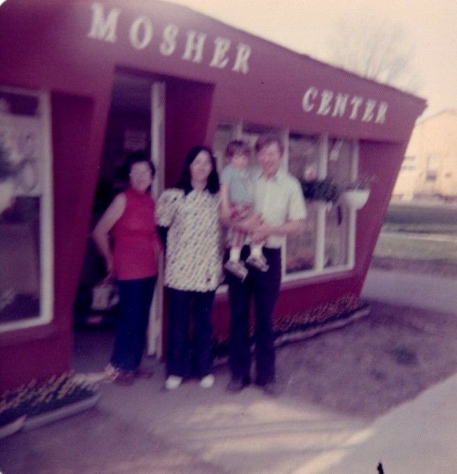 Three people standing outside a red MOSHER CENTA building, one holding a small child