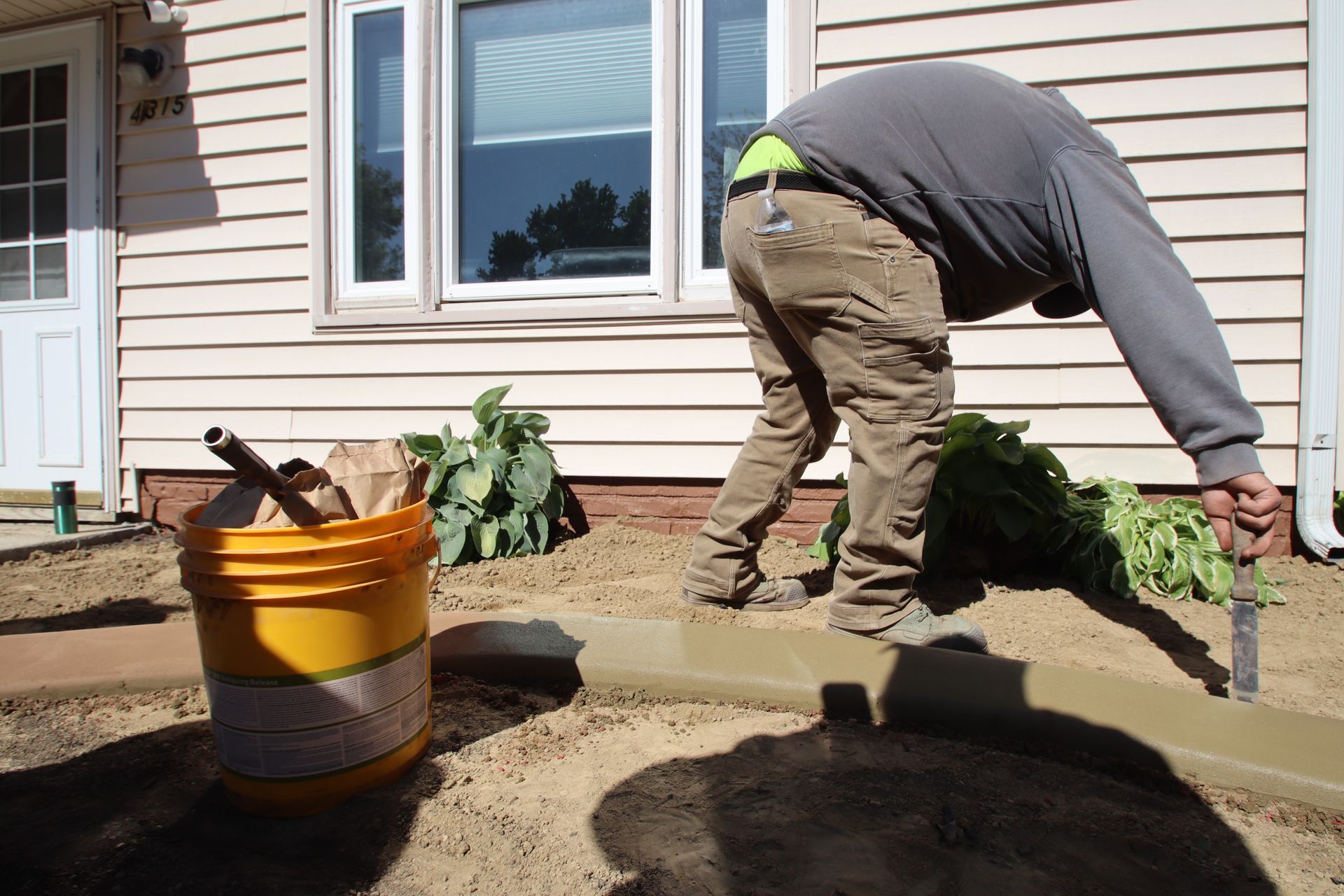 Worker pouring concrete beside a house, with a bucket and garden plants nearby.
