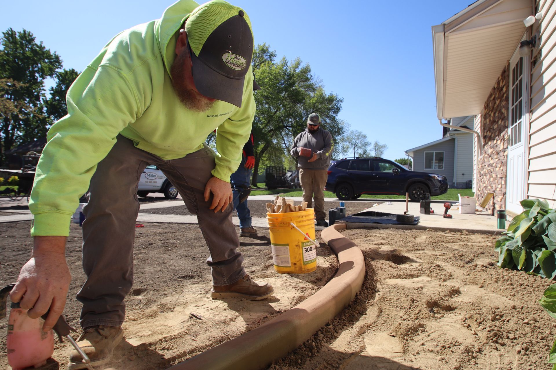 Worker in neon hoodie installs a curved garden border beside a house, with tools and mulch nearby.