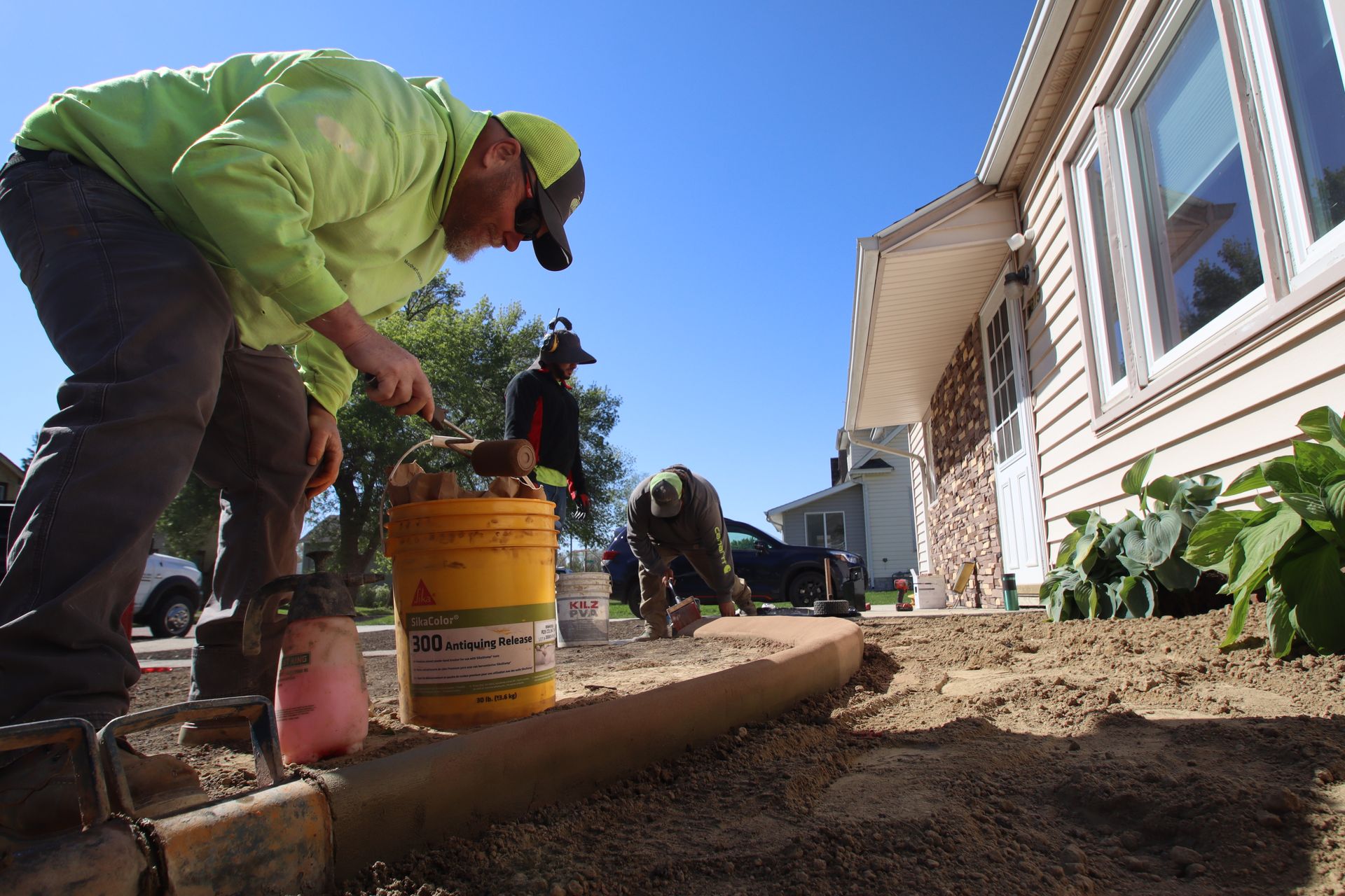 Workers landscaping beside a house, using tools and buckets in a dirt garden bed.