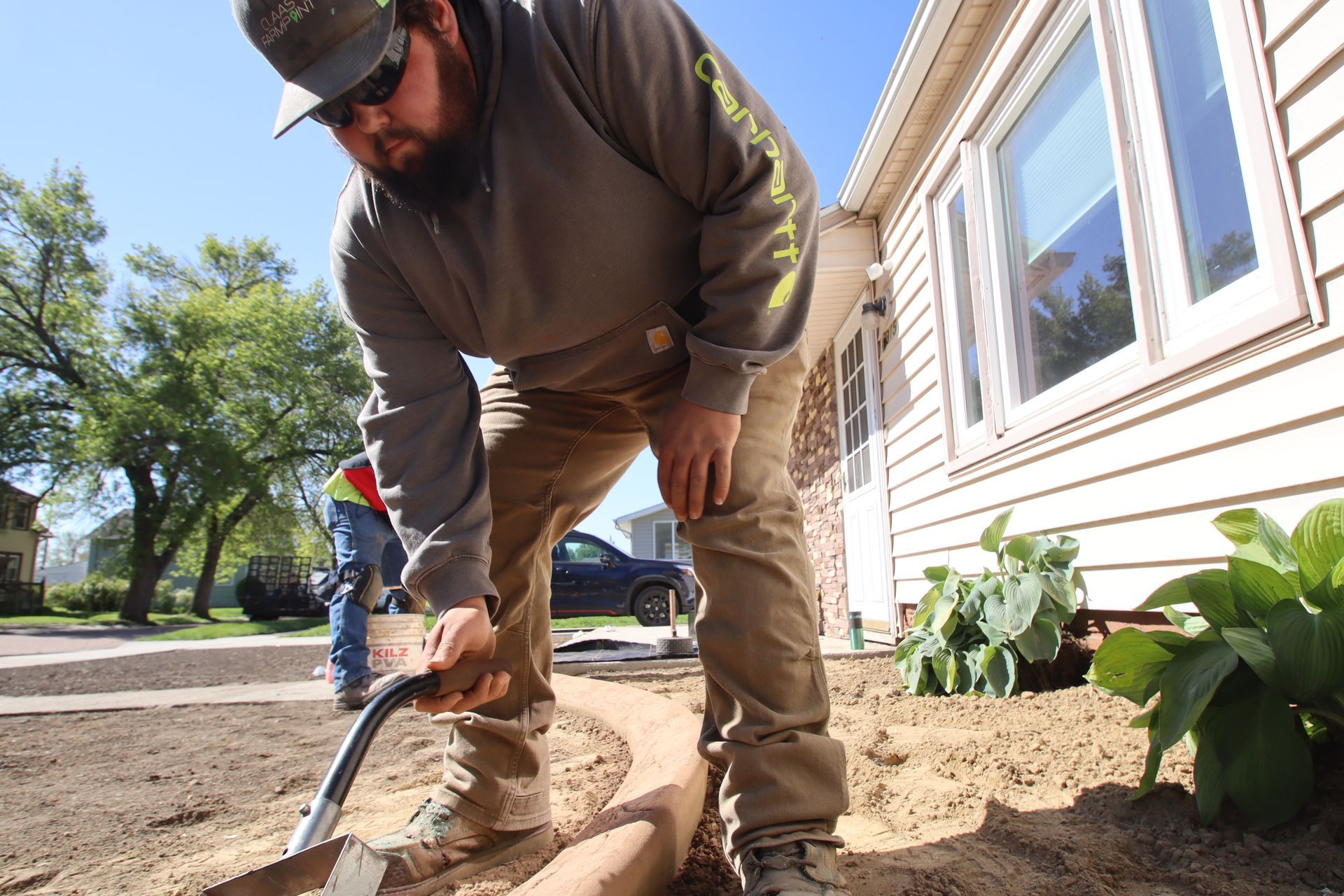Person kneeling in a garden bed, digging soil beside a house on a sunny day.