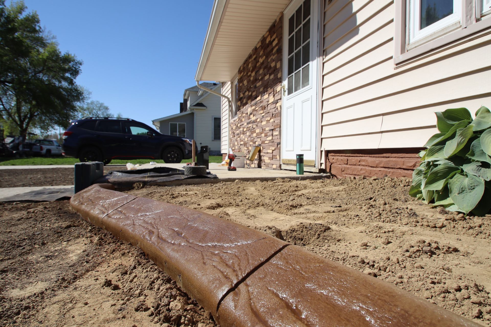 Sidewalk edge beside a house under construction, with exposed soil, a drain pipe, and a parked SUV nearby.