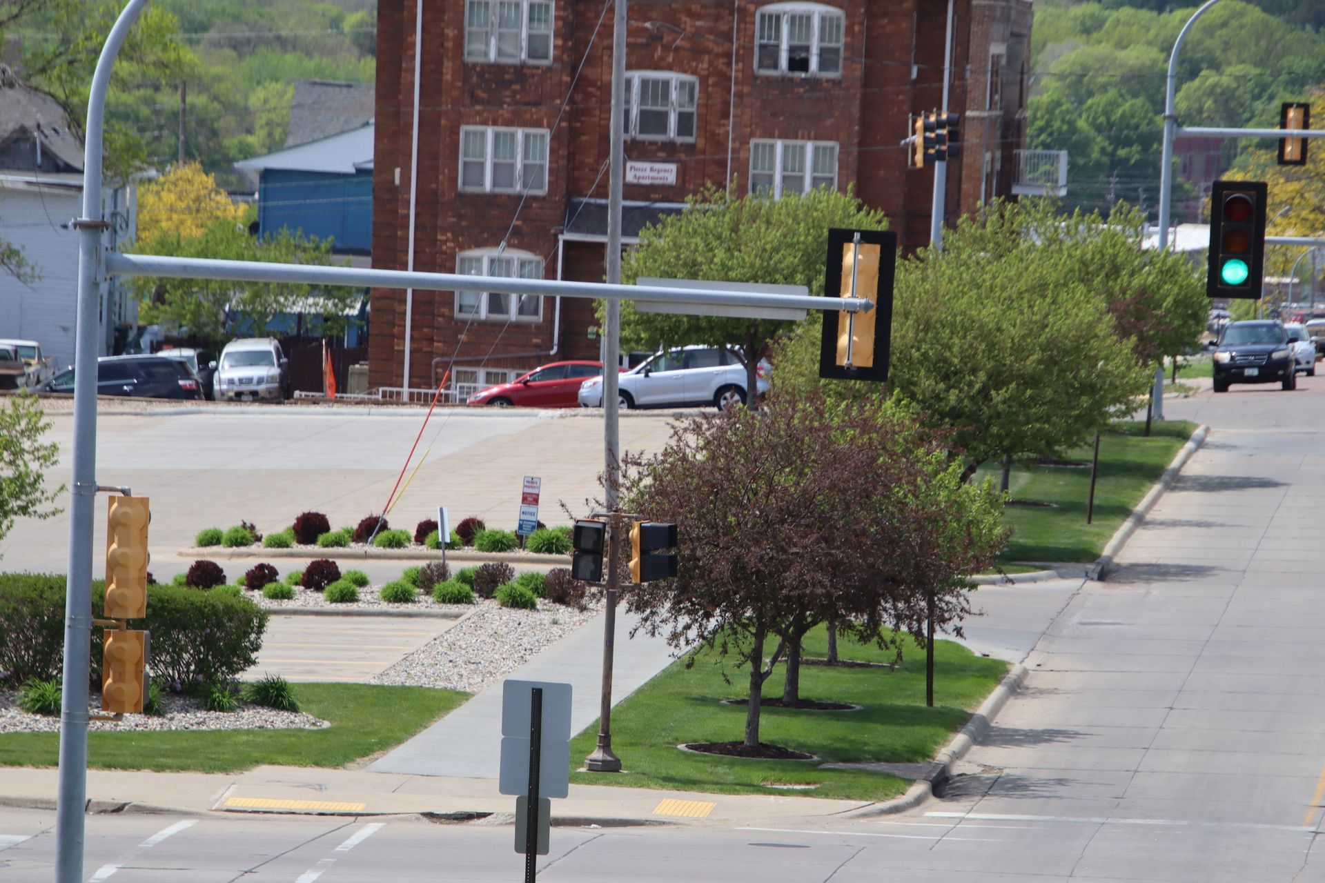 City intersection with traffic lights, cars, trees, and a brick building in the background.