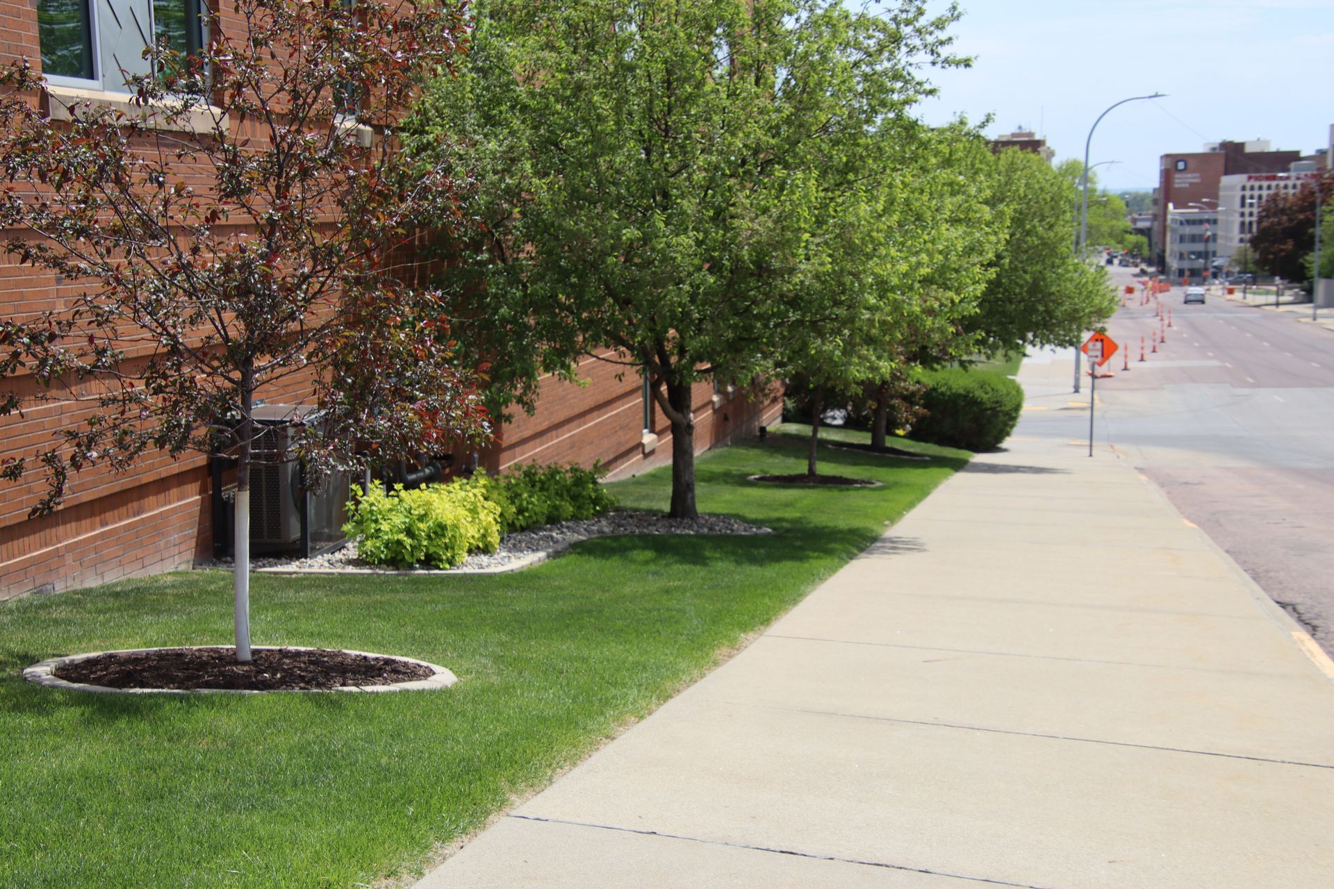 Sidewalk beside a brick building with green trees and grass on a sunny day