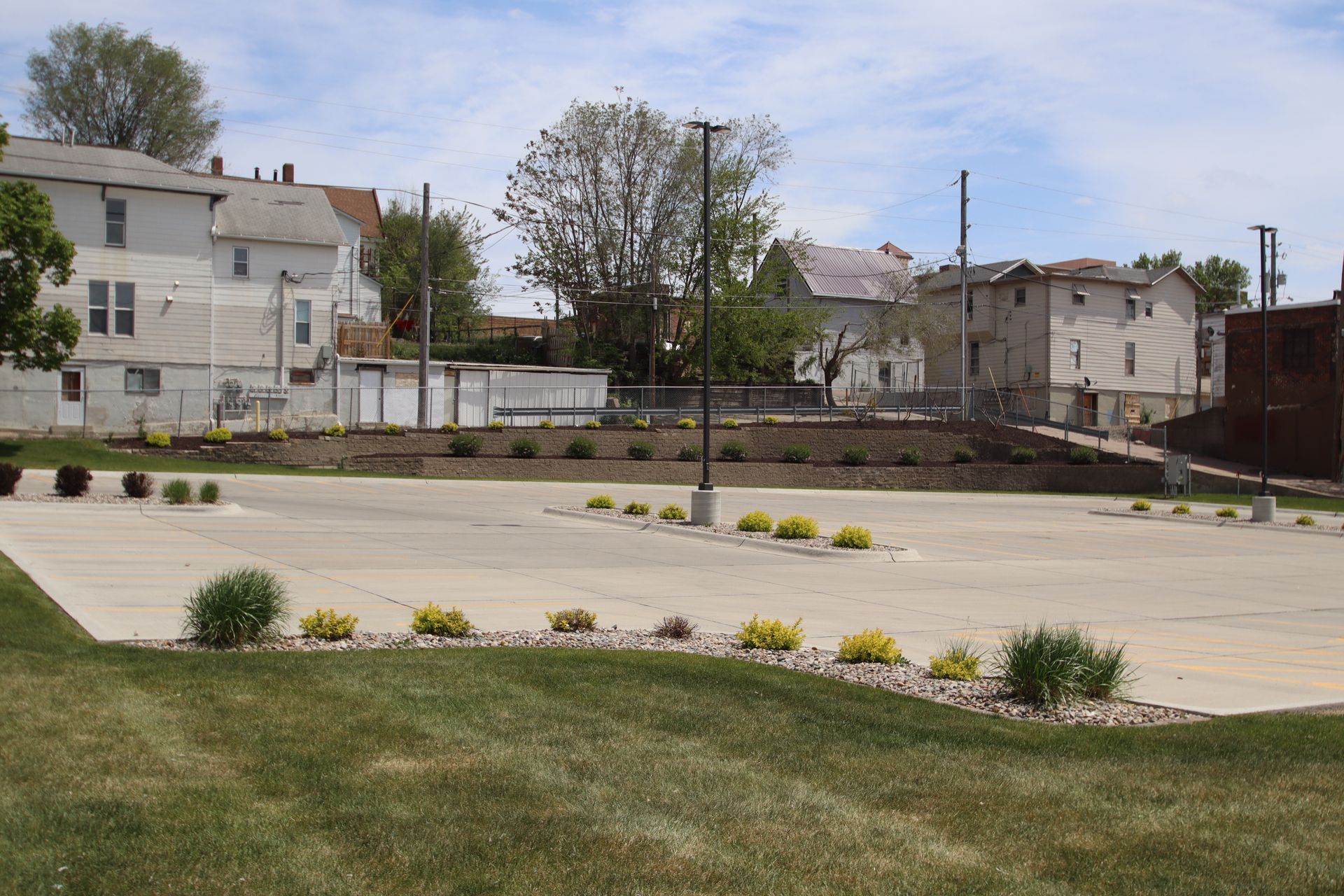 Empty paved lot with small landscaped shrubs, grass, and houses in the background