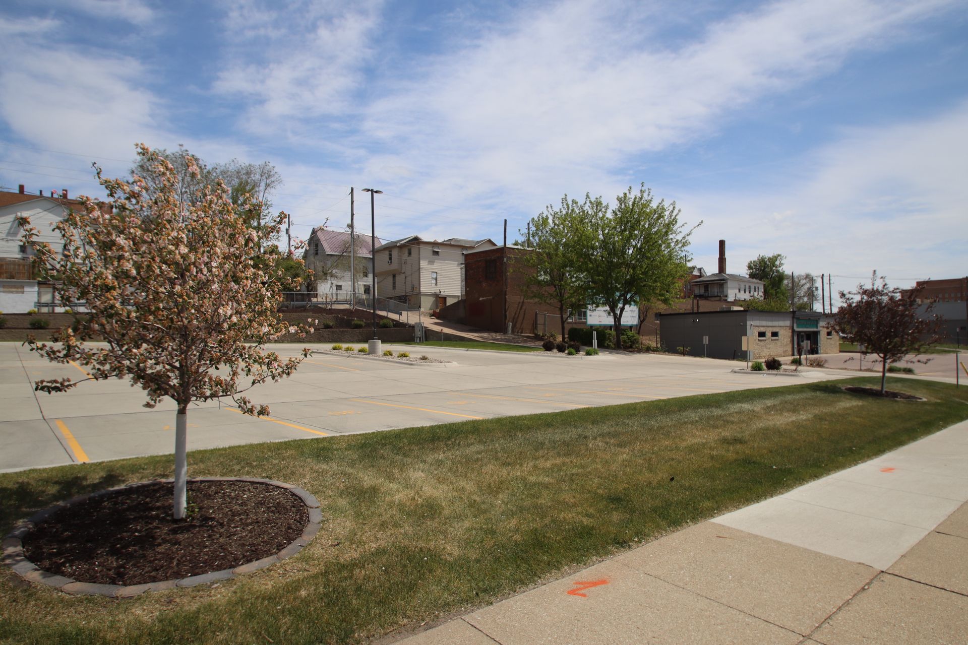 Empty parking lot beside a sidewalk and grassy strip with young trees under a cloudy sky