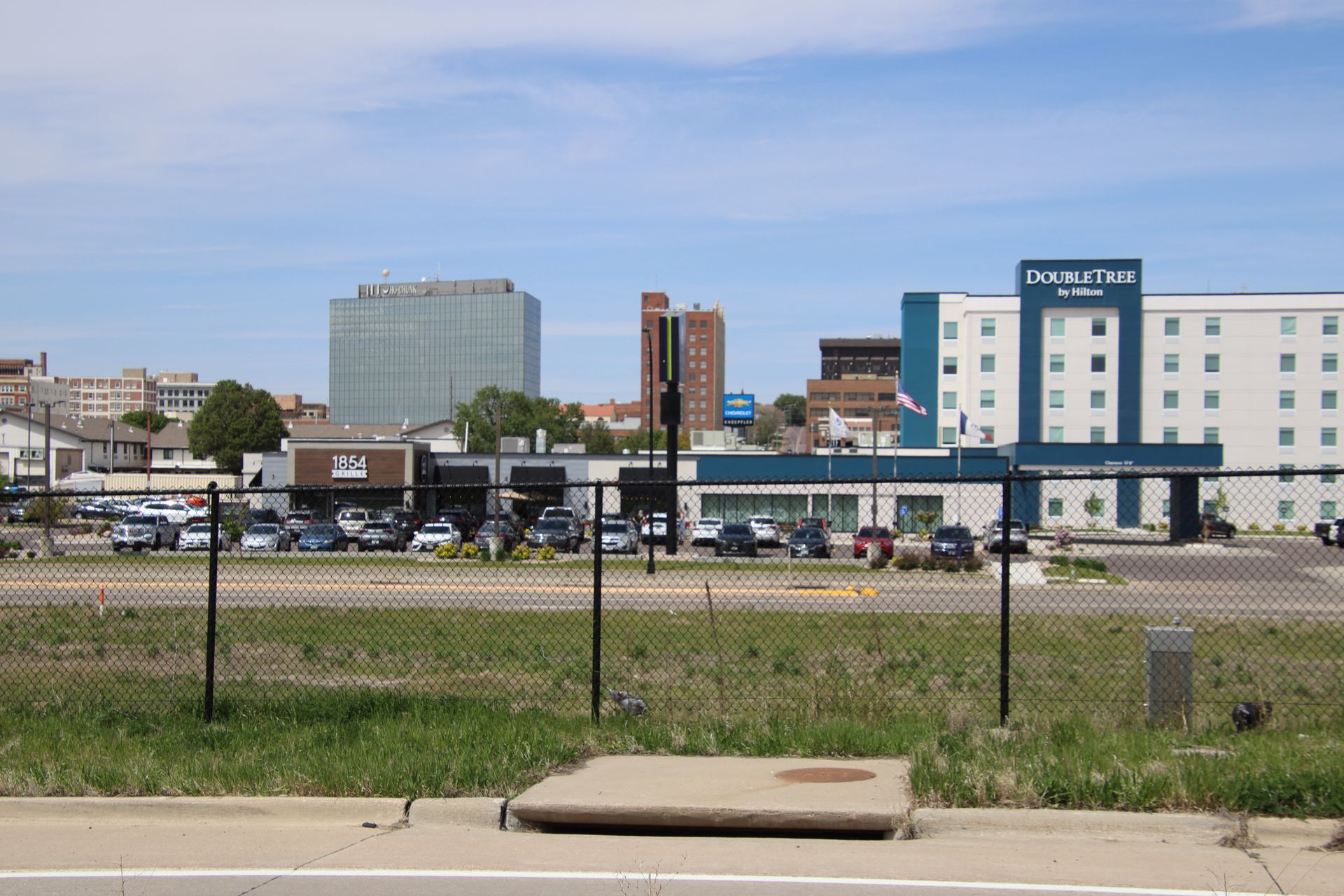 Urban street view with grassy lot, chain-link fence, and low-rise buildings under a blue sky