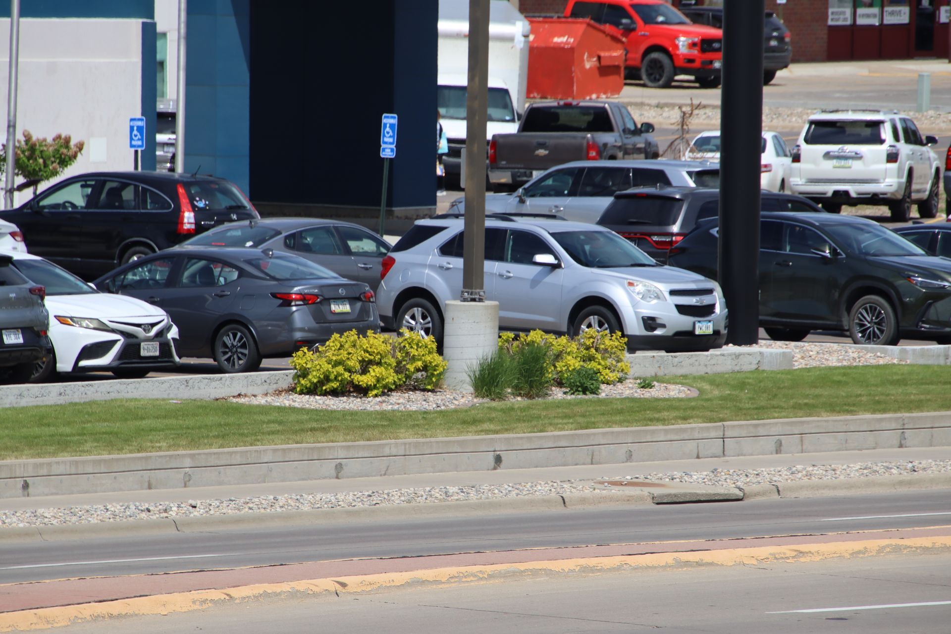 Parked cars in a lot behind a curb and roadside median with flowers and poles