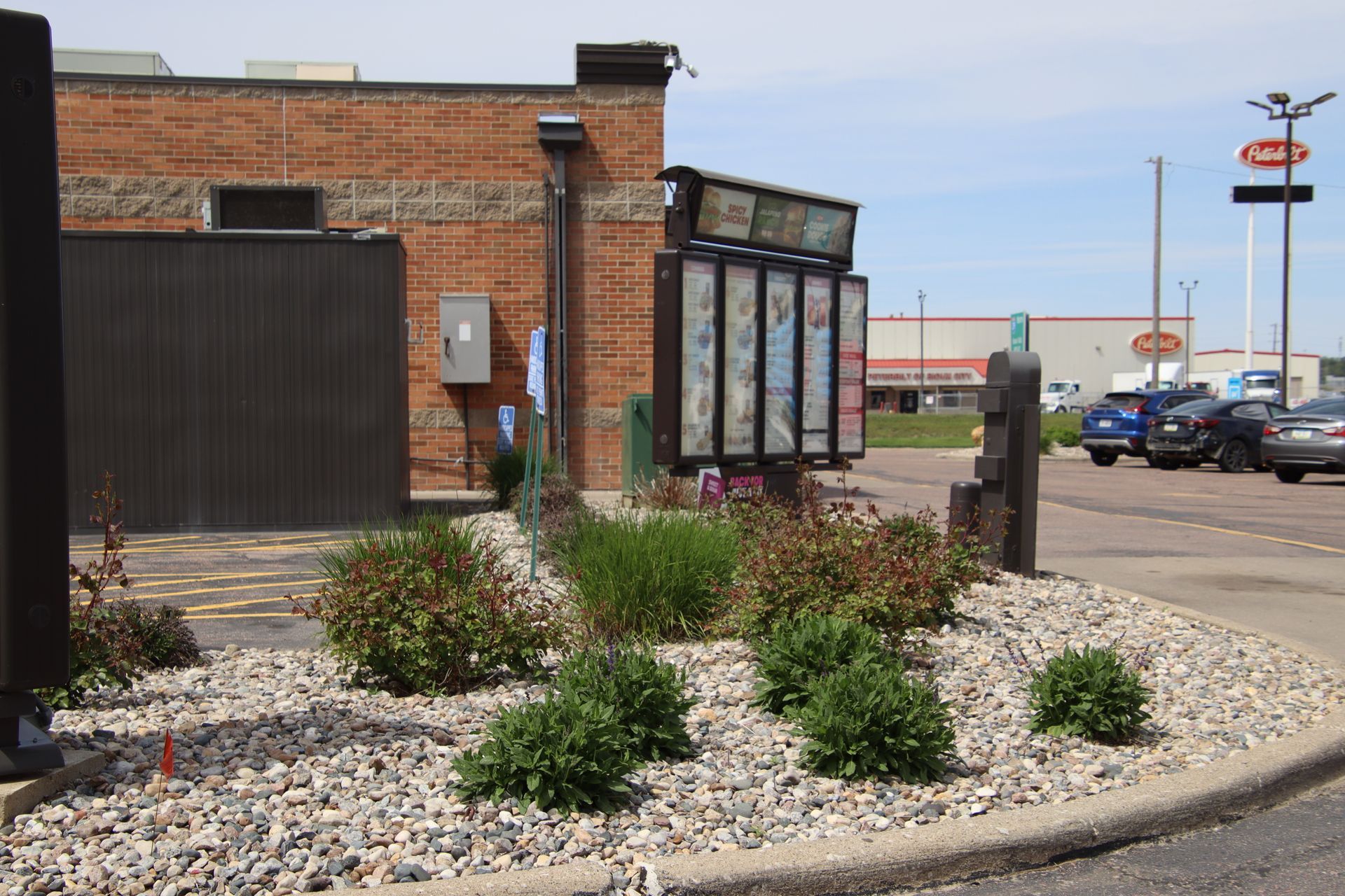 Brick storefront with gravel landscaping and a parking lot beside a road on a sunny day