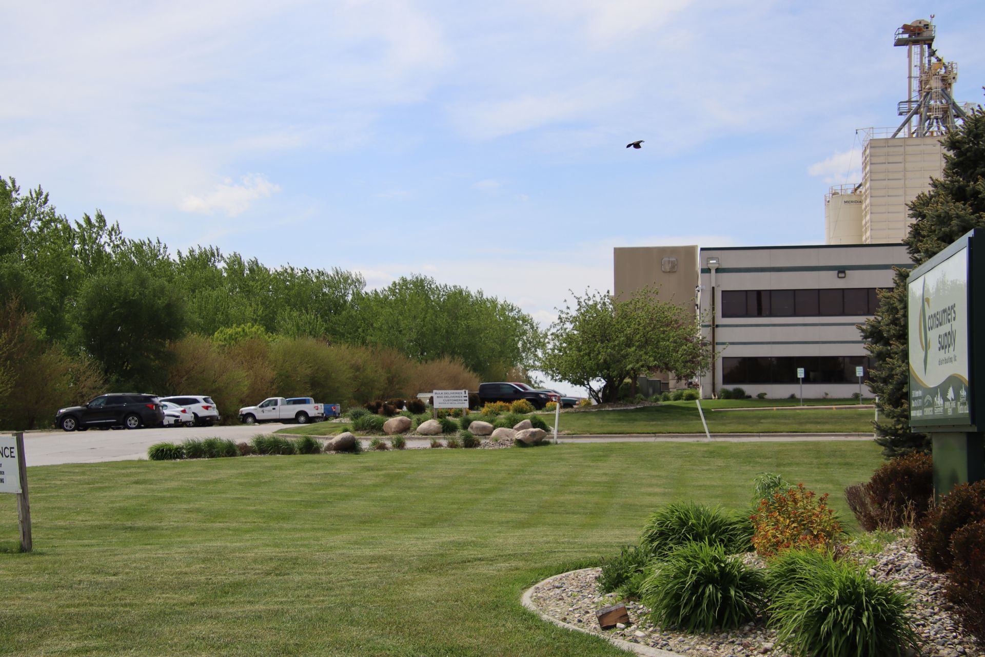 Grassy campus lawn with trees, parked cars, a building, and a tower under a partly cloudy sky