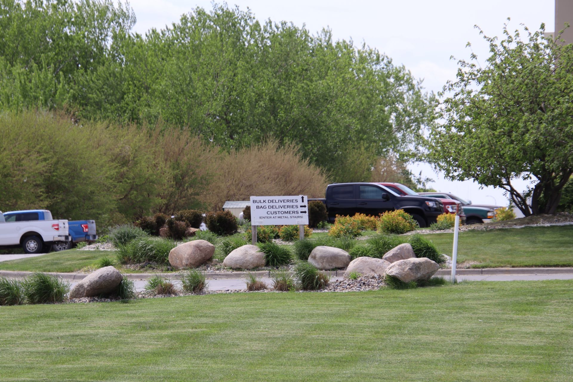 Landscaped roadside entrance with rocks, a sign, trees, and parked vehicles.