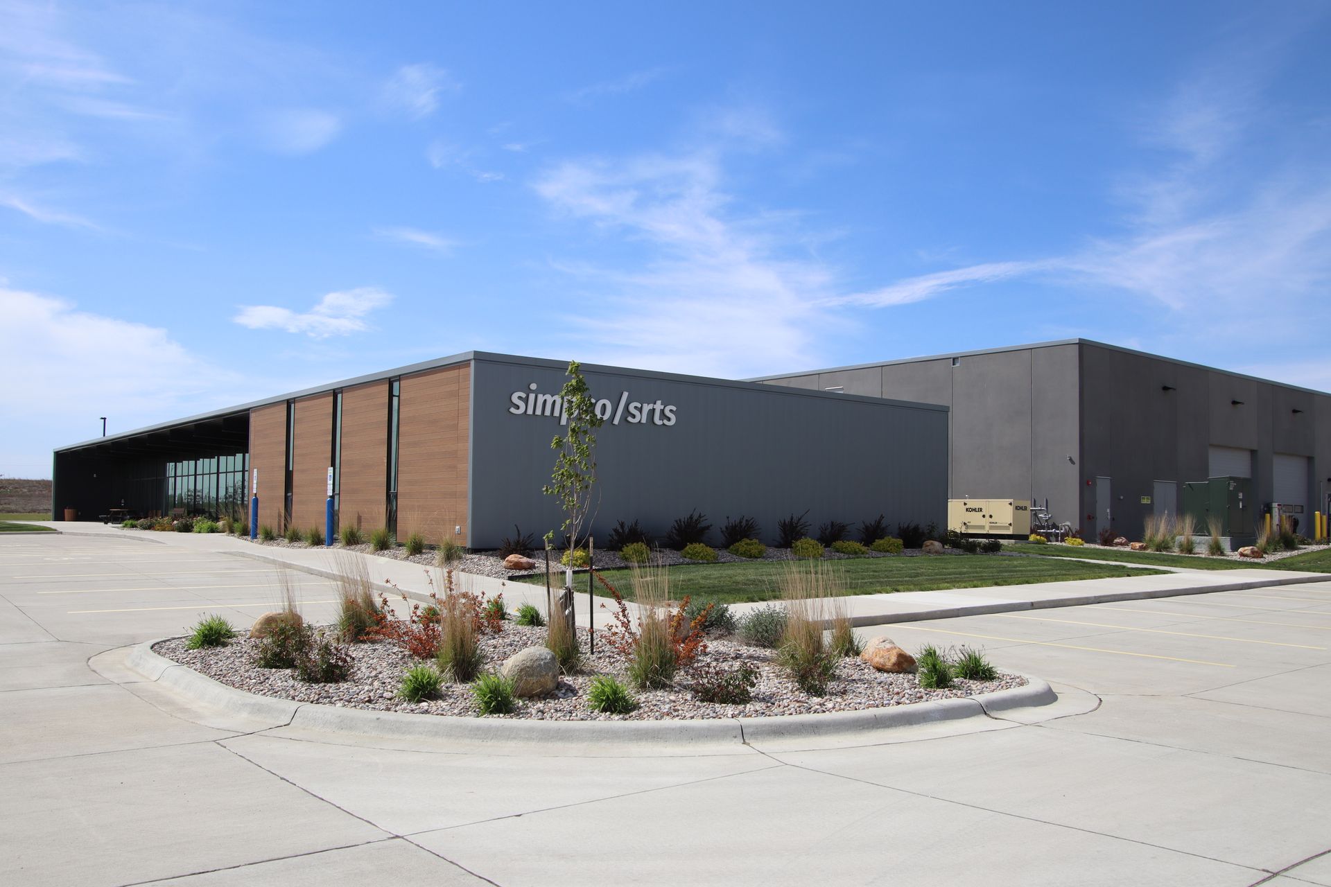 Modern industrial building with gray and tan panels, landscaped island, and blue sky above.