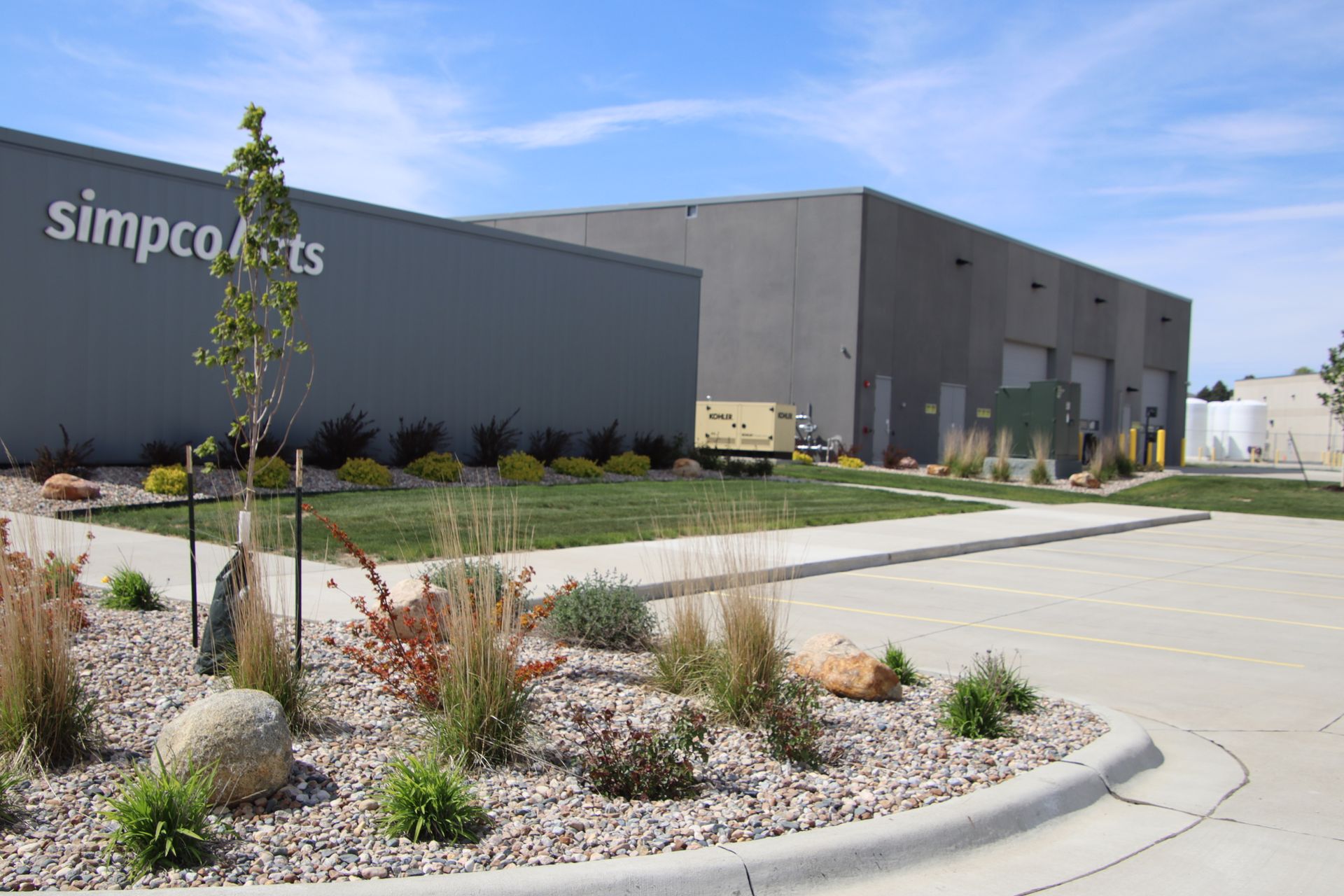 Modern industrial building with “simpco” sign, landscaped curb, and parked utility vehicles under a blue sky
